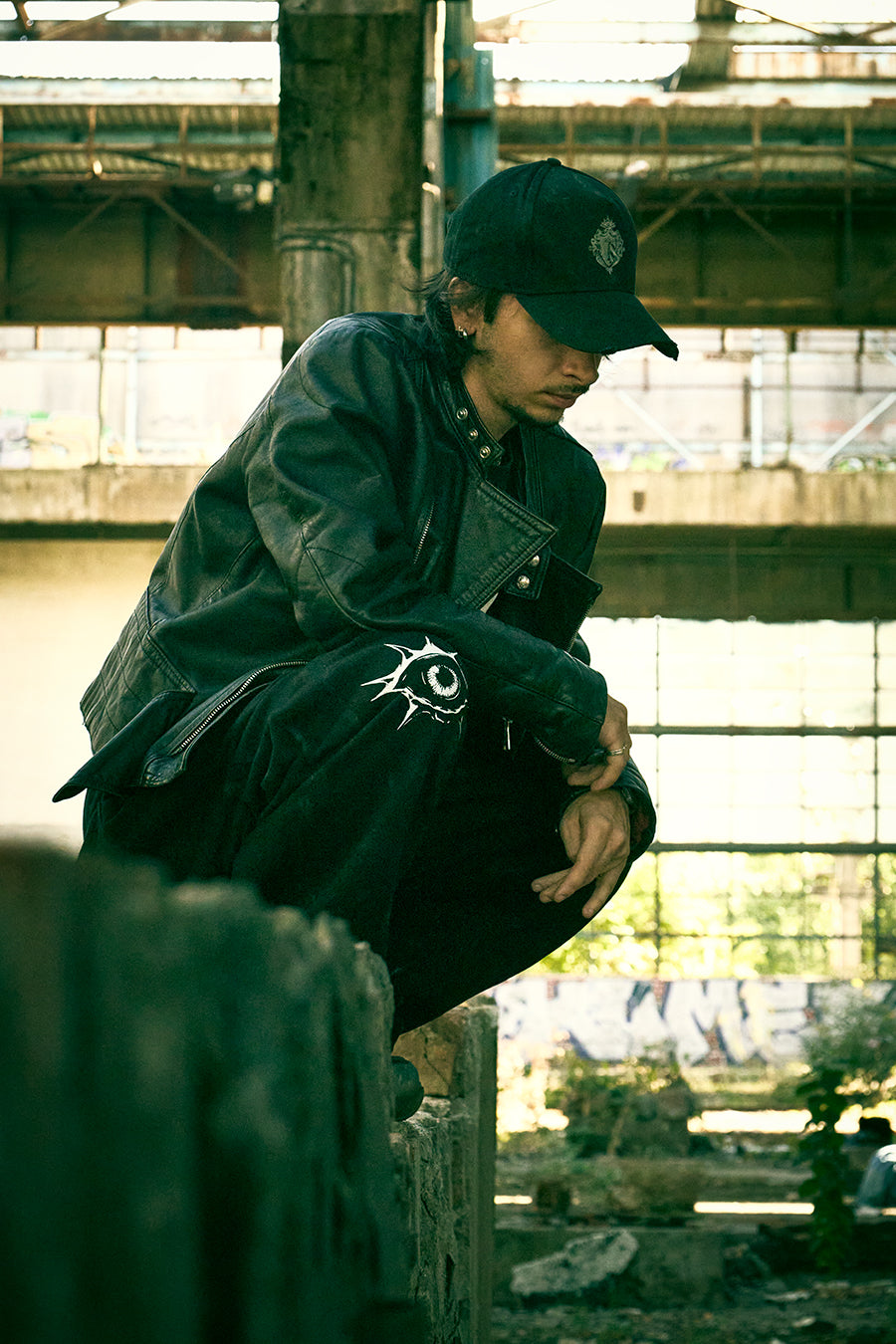 Model crouching in an abandoned industrial building, wearing the black Trips streetwear cap, a black leather jacket, and graphic print pants.